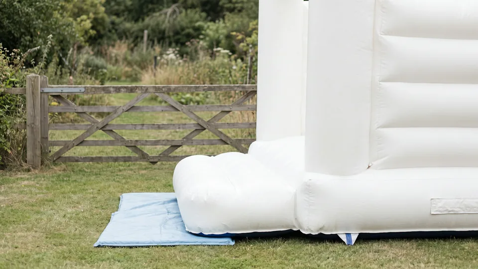 Base de um castelo inflável branco montado num gramado, com portão de madeira ao fundo e ninguém em cena
